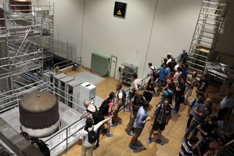 Teilnehmer*innen der Sommerschule 2016 beim Virgo-Gravitationswellendetektor nahe Pisa Eine Gruppe von Menschen in einem lagerhallenartigen Raum mit Gerüst und mehreren großen Geräten