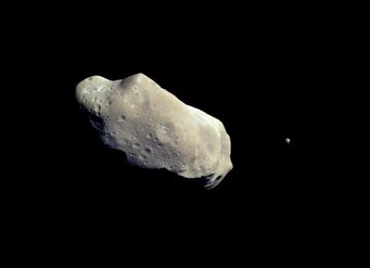 A potato-shaped, gray-brown object with a cratered surface and a small moon on a black background A potato-shaped, gray-brown object with a cratered surface and a small moon on a black background