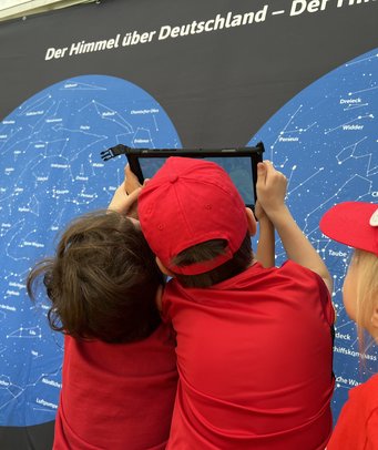 Forscheraufgabe: Vor welchem Sternbild befindet sich gerade die Sonne? Mit Hilfe der App lässt sich das live herausfinden! Zu sehen sind zwei Grundschulkinder von hinten, die vor zwei senkrechten Sternbildkarten stehen. Sie blicken auf ein Tablet, dass sie gemeinsam hoch in den Himmel halten.