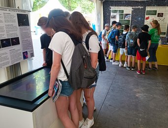 Mitmachen beim Citizen-Science-Projekt Quallen-Galaxien Eine Gruppe Teenager vor einem Touchtable, vor ihnen hängt ein Poster, das das Quallengalaxien-Projekt erklärt. Im Hintergrund eine Gruppe jüngerer Kinder, die einer Standbetreuerin zuhört