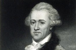 Schwarzbild eines Mannes mit lockigen Haaren hält ein Papier mit einer Zeichnung des Planeten Uranus mit zwei Monden. Er trägt ein weißes Hemd, darüber einen Mantel mit flauschigem Kragen.