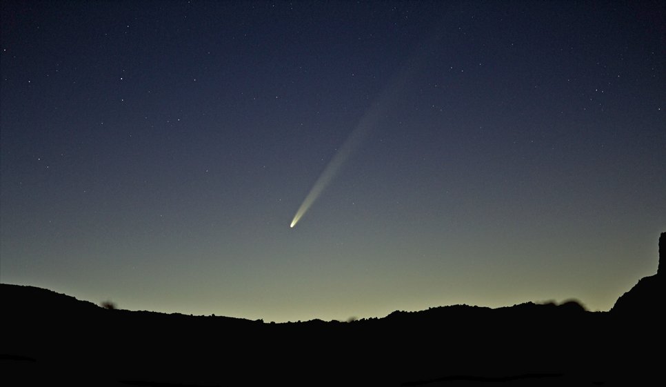 A comet with clearly distiguishable tail above the silhouette of a landscape without much detail at dawn
