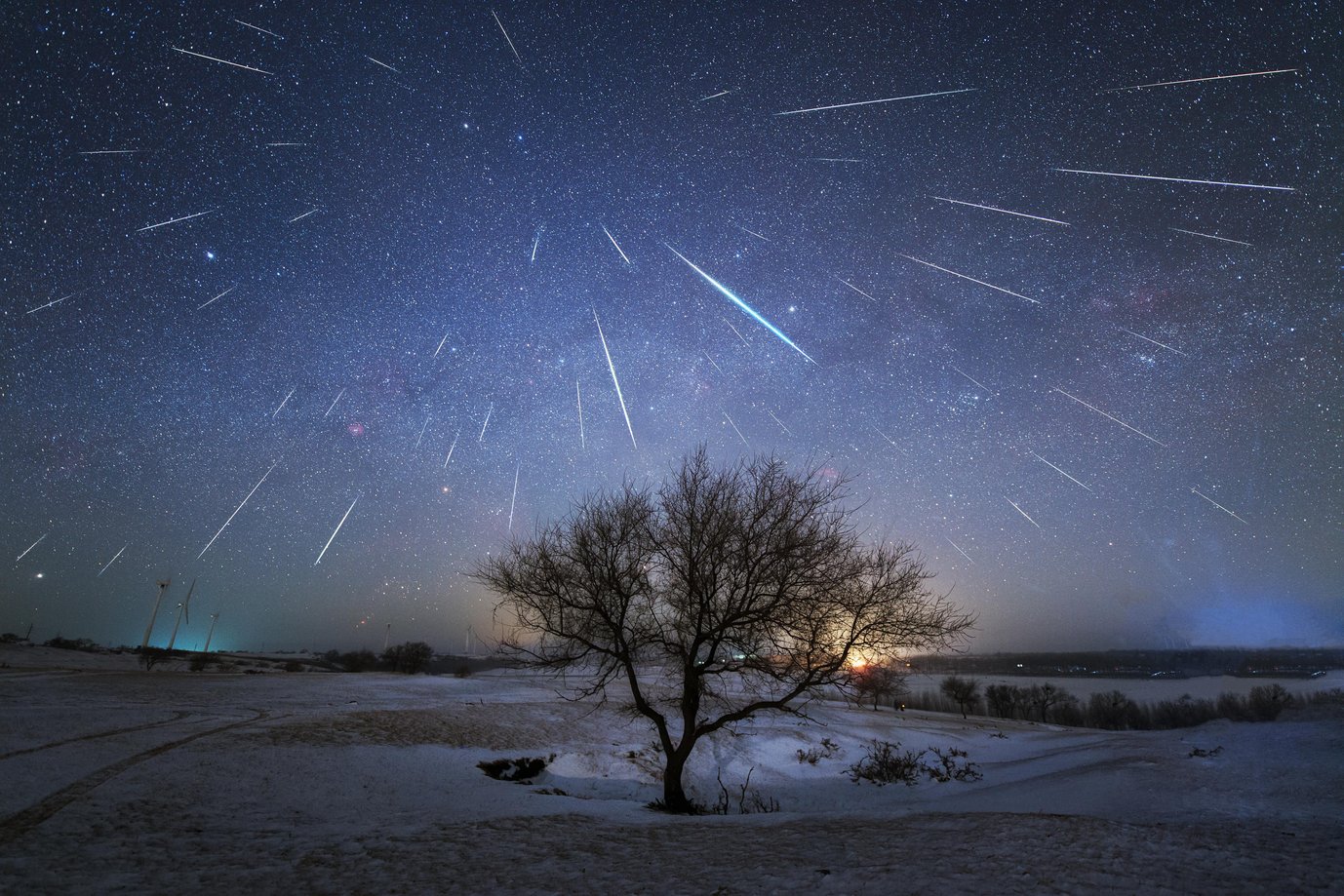 Countless shooting stars, some of them with a greenish color, spreading out in all directions from a point in the starry sky above a snow-covered landscape with a bare tree in the center of the image.