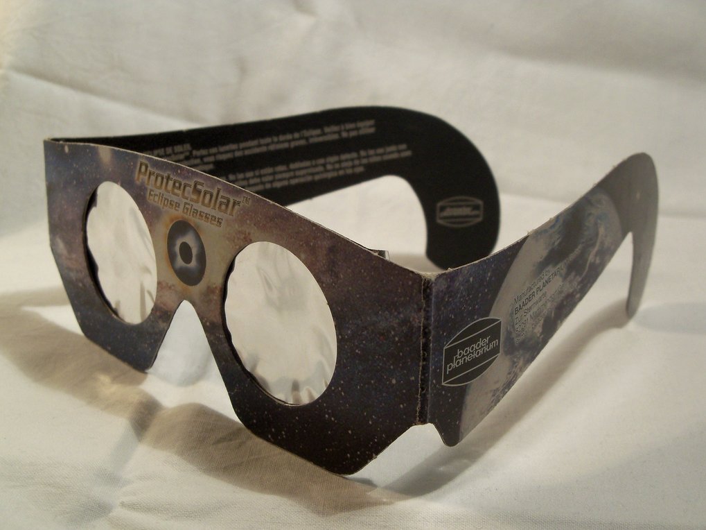 Sonnenfinsternisbrille aus Pappe mit zwei runden Sichtfenstern aus reflektierender Schutzfolie. Die Brille ist dunkel bedruckt und zeigt ein Weltraummotiv mit Sternen und Planeten. Auf dem linken Bügel befindet sich ein rechteckiges Etikett mit weißer Schrift. Die Brille steht aufgeklappt auf einer hellen, glatten Oberfläche.