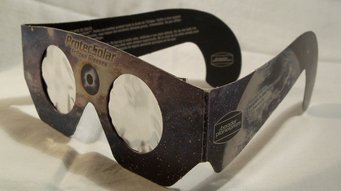 Sonnenfinsternisbrille mit spezieller reflektierender Folie, die eine gefahrlose Beobachtung der Sonne ermöglicht. Sonnenfinsternisbrille aus Pappe mit zwei runden Sichtfenstern aus reflektierender Schutzfolie. Die Brille ist dunkel bedruckt und zeigt ein Weltraummotiv mit Sternen und Planeten. Auf dem linken Bügel befindet sich ein rechteckiges Etikett mit weißer Schrift. Die Brille steht aufgeklappt auf einer hellen, glatten Oberfläche.