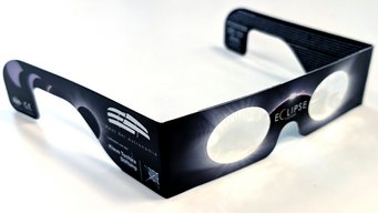 Sonnenfinsternisbrille mit spezieller reflektierender Folie, die eine gefahrlose Beobachtung der Sonne ermöglicht. Sonnenfinsternisbrille aus Karton mit zwei runden Sichtfenstern aus reflektierender Schutzfolie. Die Brillenbügel sind breit und tragen weiße Aufdrucke, darunter ein Logo, Schriftzüge und ein Piktogramm. Die Vorderseite zeigt eine grafische Darstellung einer Sonnenfinsternis mit leuchtendem Rand um die Sichtfenster. Die Oberfläche wirkt matt, die Schutzfolie glänzt leicht.