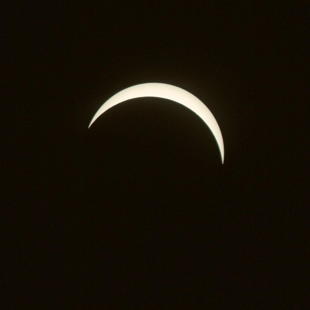 Eine helle, schmale Sichel steht vor dunklem Hintergrund. Die Form zeigt eine partielle Sonnenfinsternis, bei der der Mond einen Teil der Sonne bedeckt. Die Konturen sind klar und gleichmäßig.