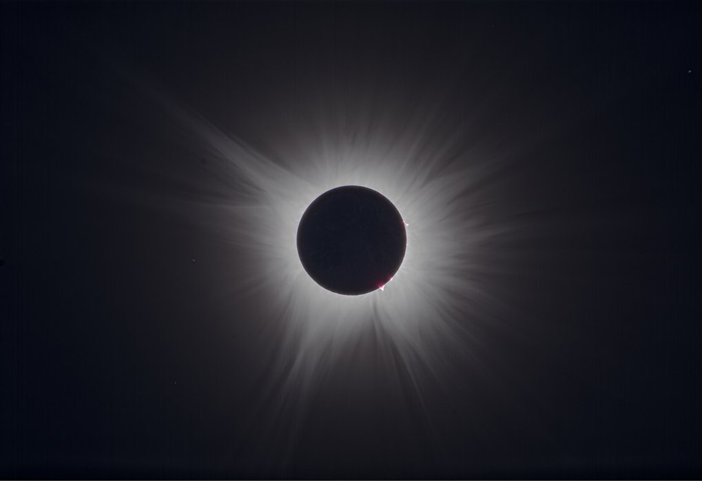 Im Zentrum steht die vollständig vom Mond bedeckte Sonne während einer Sonnenfinsternis. Die helle Korona der Sonne bildet einen strahlenförmigen, unregelmäßigen Lichtkranz um die dunkle Mondscheibe. An mehreren Stellen am Sonnenrand sind rötliche Ausbrüche sichtbar, die als Protuberanzen bezeichnet werden. Der Hintergrund ist dunkel, wodurch die feinen Strukturen der Sonnenkorona deutlich hervortreten.